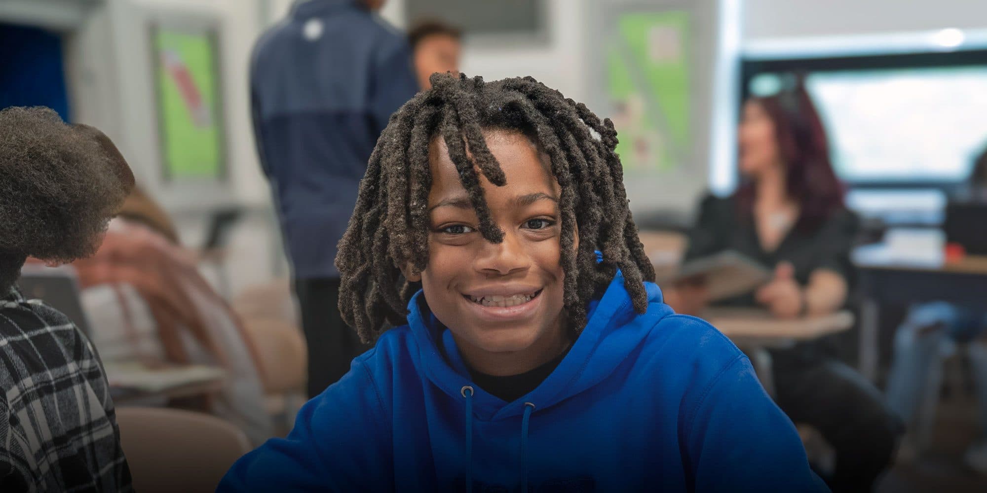 Smiling student in a blue hoodie sitting in a classroom.