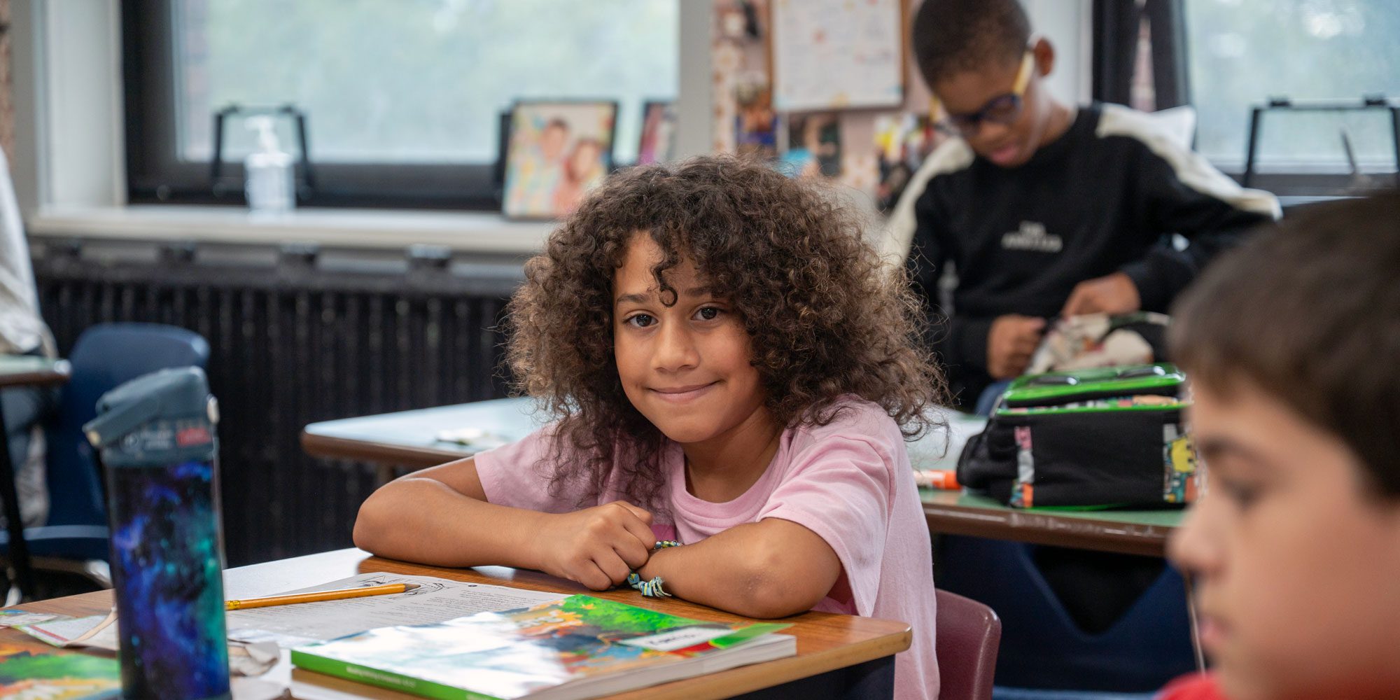Smiling student with curly hair seated at a classroom desk with books.
