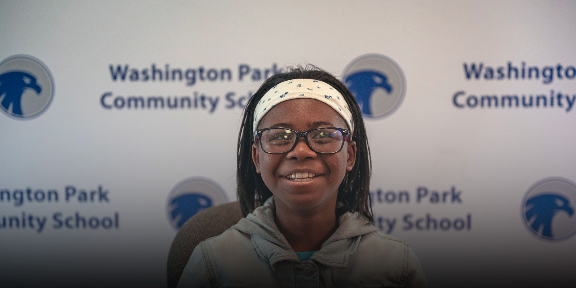Smiling student with glasses in front of a Washington Park Community School backdrop.