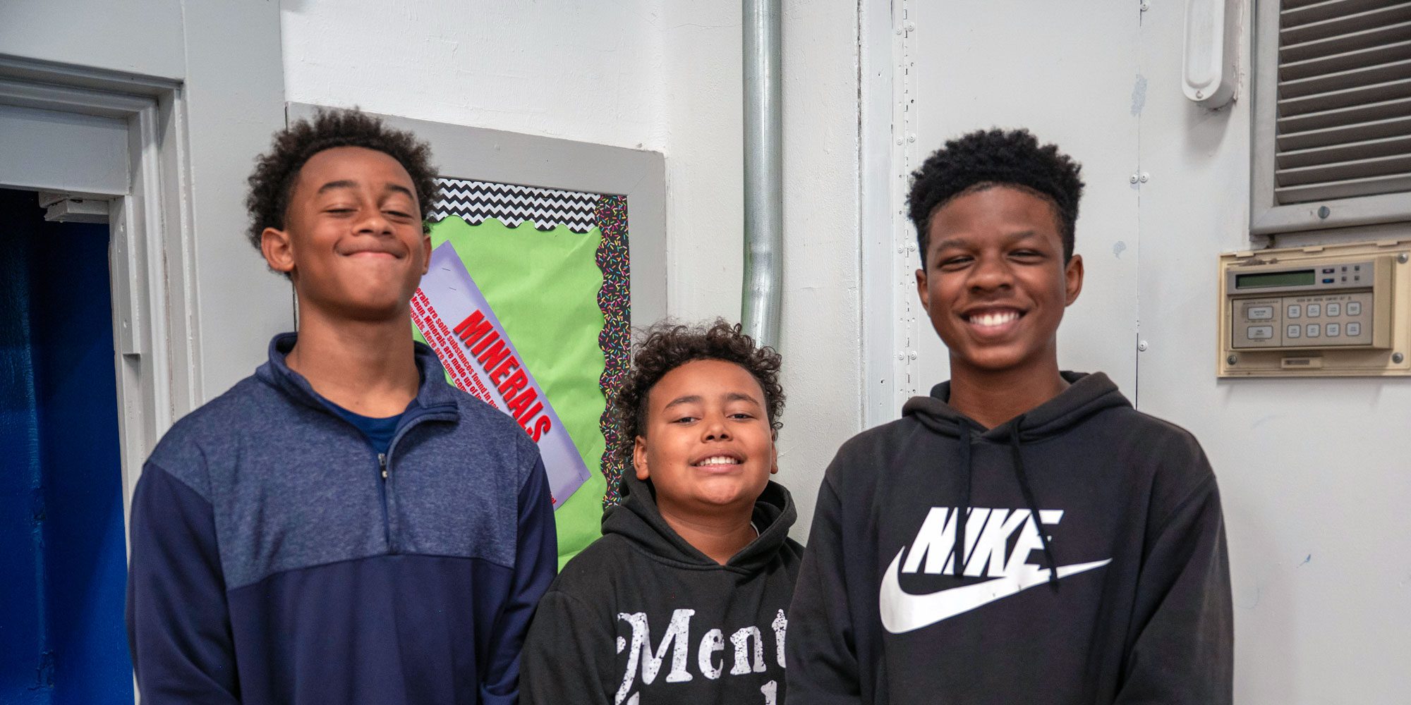 Three smiling students standing together in front of a classroom bulletin board.